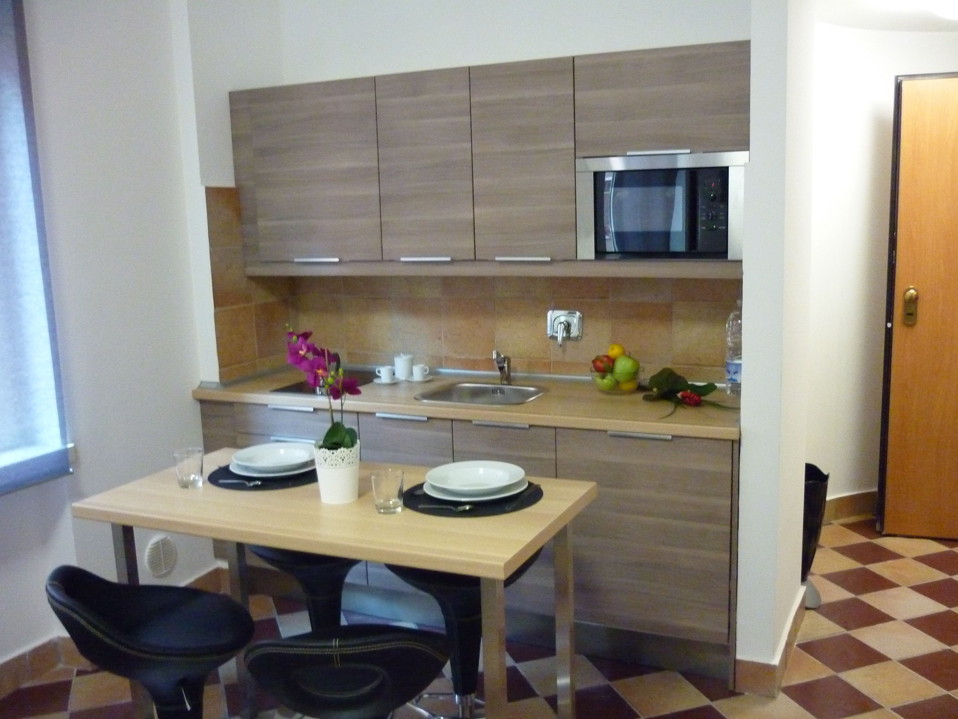 A compact kitchen with a microwave, sink, and a small fruit bowl on the counter. There is a table with two chairs and a vase with flowers.