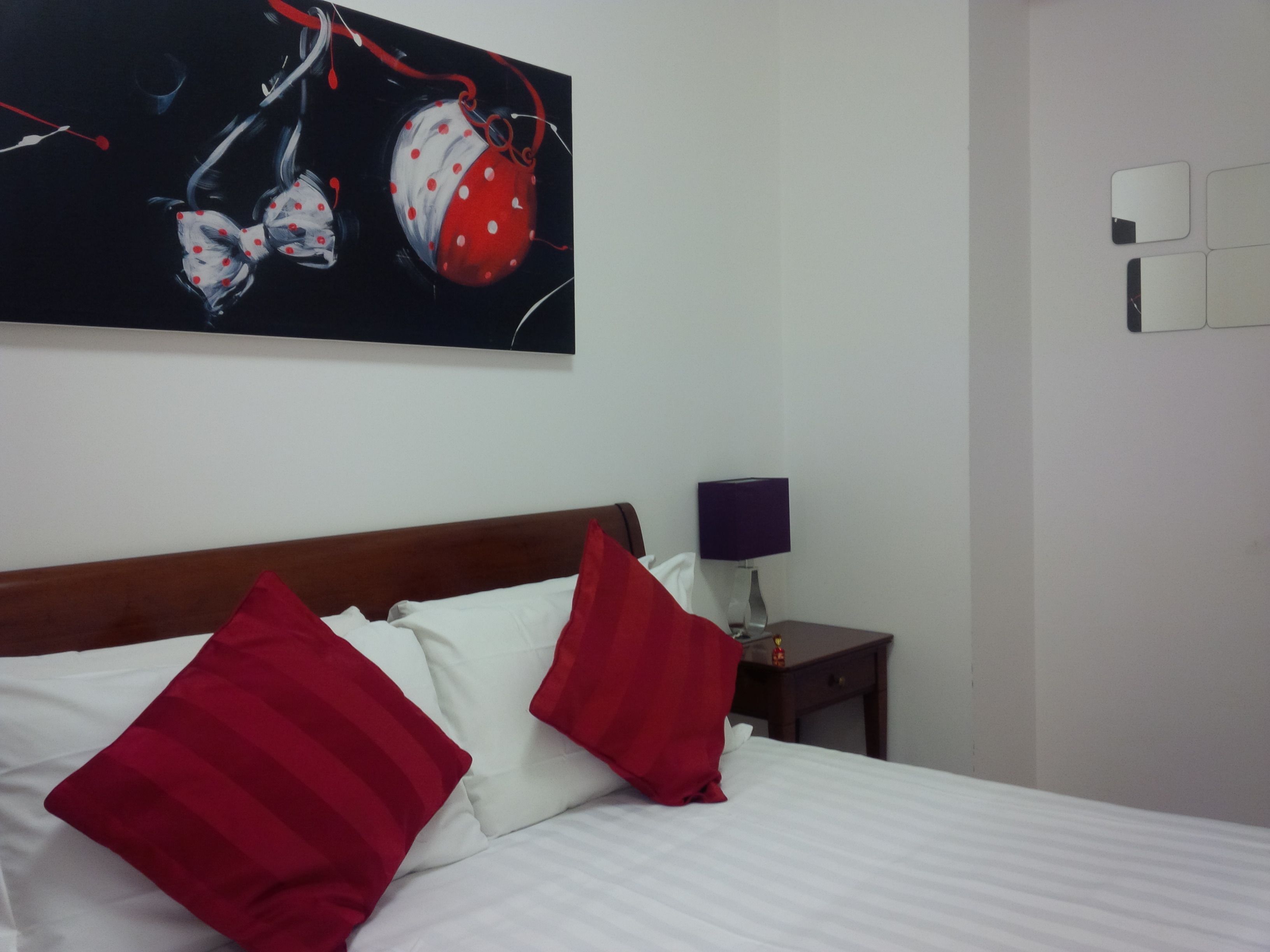 Una camera d'albergo con un letto, cuscini rossi e bianchi, un quadro sopra il letto, un comodino con una lampada e uno specchio fissato al muro.