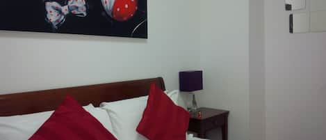 Una camera d'albergo con un letto, cuscini rossi e bianchi, un quadro sopra il letto, un comodino con una lampada e uno specchio fissato al muro.