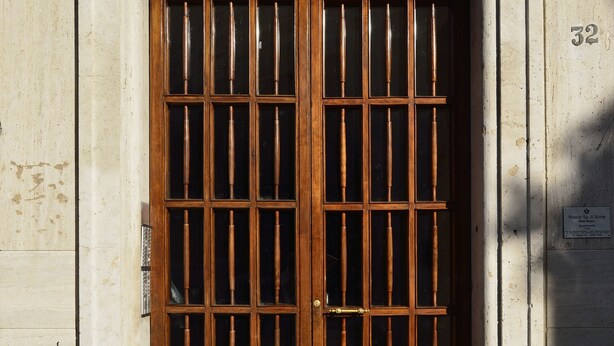 A large wooden door with vertical bars, set in a stone wall with a number plaque. - Maison Re di Roma (Rome)