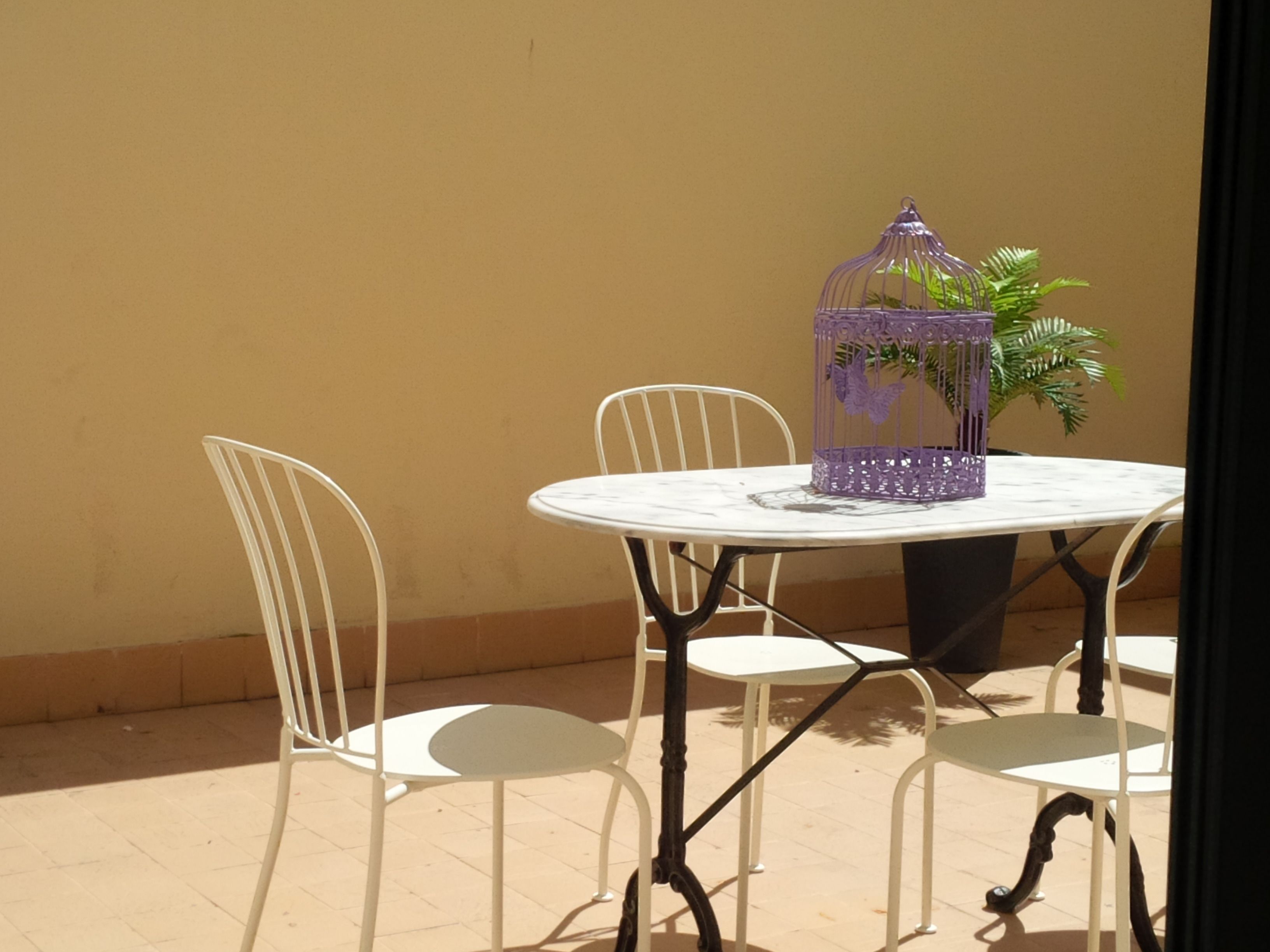 A round table with a white marble top, surrounded by four white metal chairs with curved backs. A purple birdcage with a plant inside is placed on the table.