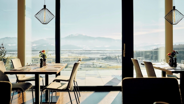 A dining area with a view of a cityscape and mountains. Tables are set with chairs, and there are hanging light fixtures. - TAUROA Schönberghof Spielberg (Spielberg)