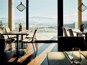Un espace repas offrant une vue sur le paysage urbain et les montagnes. Des tables sont disposées avec des chaises, et des luminaires sont suspendus au plafond.