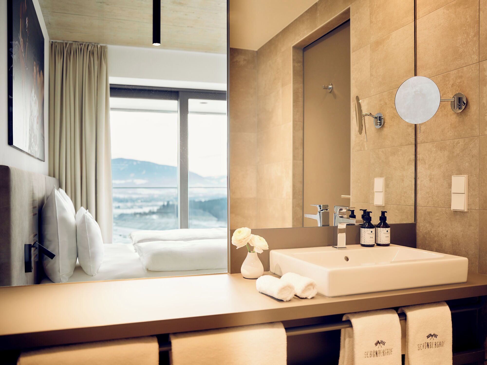 A modern bathroom with a large mirror, a sink, and toiletries. There is a window with a view of mountains.