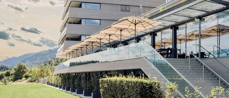 Un bâtiment moderne à plusieurs étages, doté d’une façade en verre, d’un espace extérieur aménagé avec des parasols et d’un escalier menant à l’entrée.
