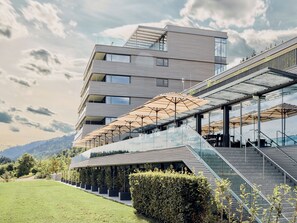 Ein modernes, mehrstöckiges Gebäude mit Glasfassade, einer Sitzterrasse mit Sonnenschirmen und einer Treppe zum Eingang.