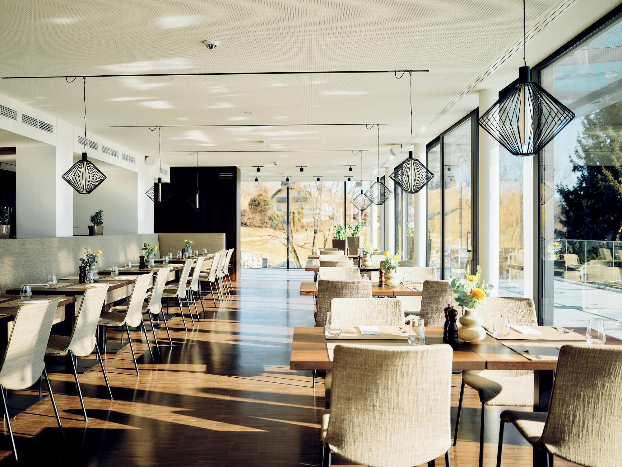 A modern dining area with long tables and chairs, large windows, and geometric pendant lighting.