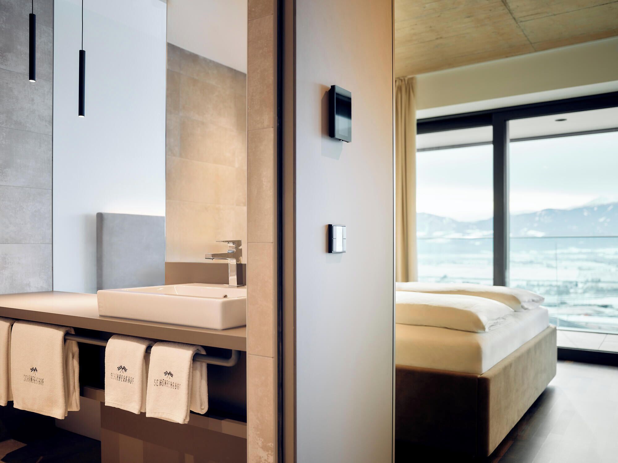 A modern bathroom with a large mirror, a sink, and a toilet. There are towels hanging on a rack. The room has a large window with a view of mountains.