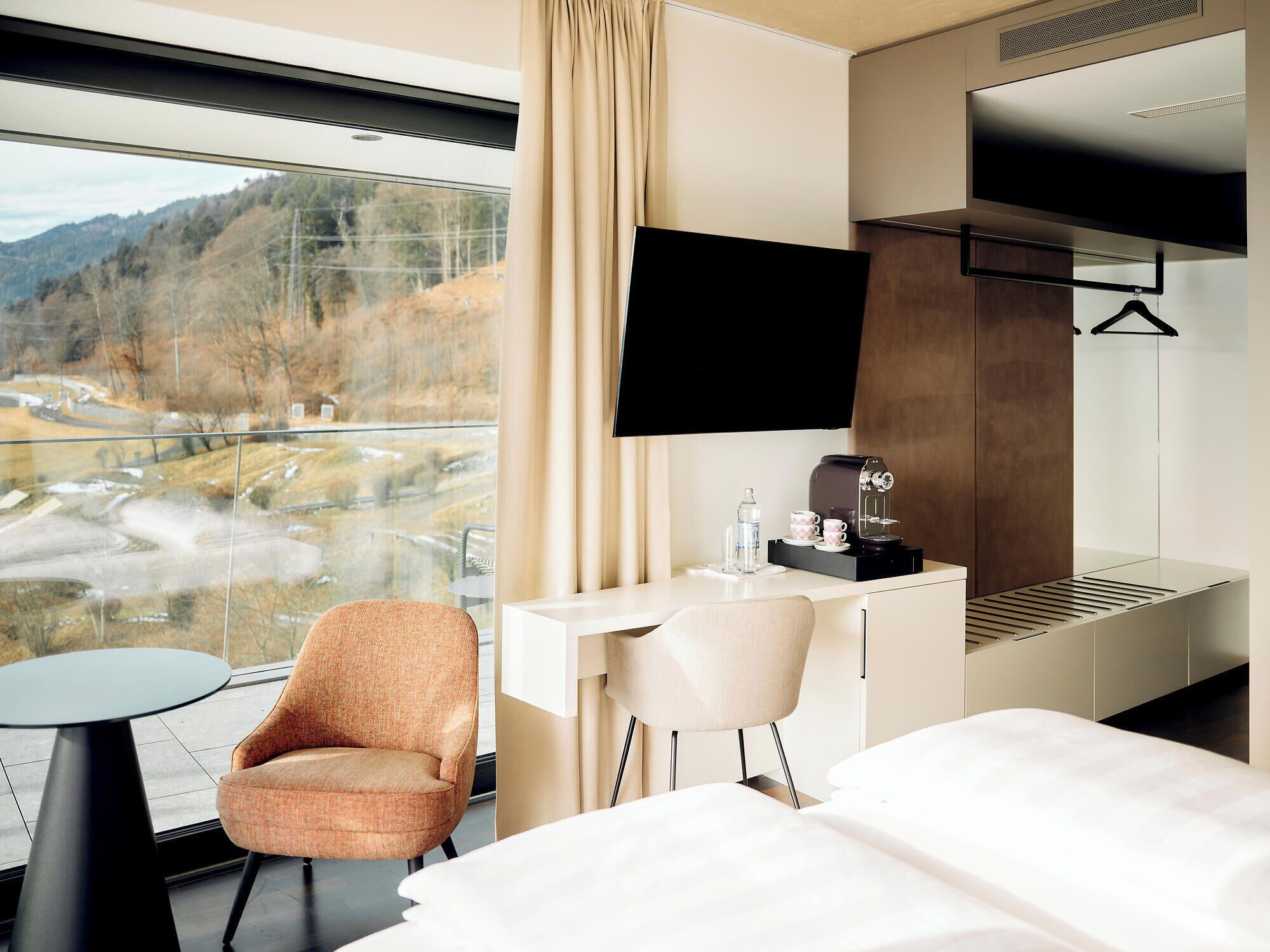 Ein Hotelzimmer mit großem Fenster und Panoramablick auf ein Tal, einem Bett, einem Schreibtisch mit Kaffeemaschine und einem an der Wand befestigten Fernseher.