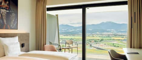 Habitación de hotel con una cama grande, un escritorio, una silla y vistas a un paisaje a través de un ventanal amplio.