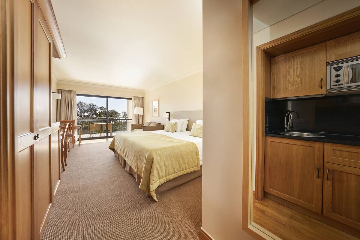 Una habitación de hotel con una cama grande, un balcón con vistas, armarios de madera y una kitchenette.