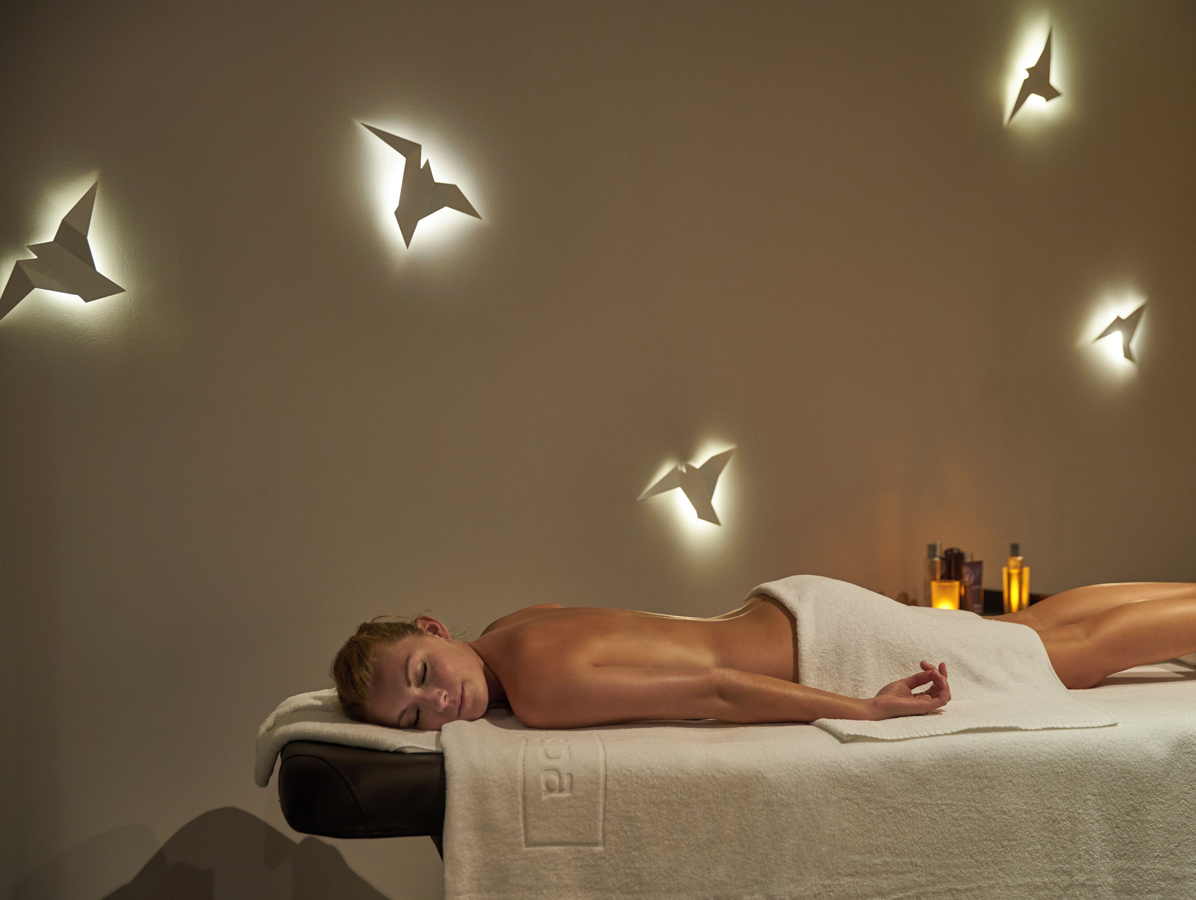 A spa setting with a person lying on a treatment table, surrounded by wall-mounted light fixtures shaped like birds.