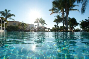 A clear swimming pool surrounded by palm trees and a building with a covered area.