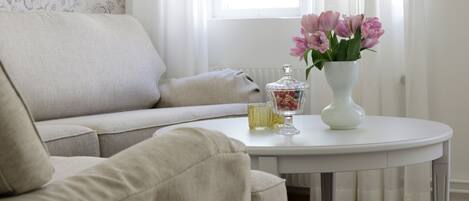Una sala de estar con un sofá beige, una mesa blanca con un jarrón de flores rosas y un recipiente de vidrio.