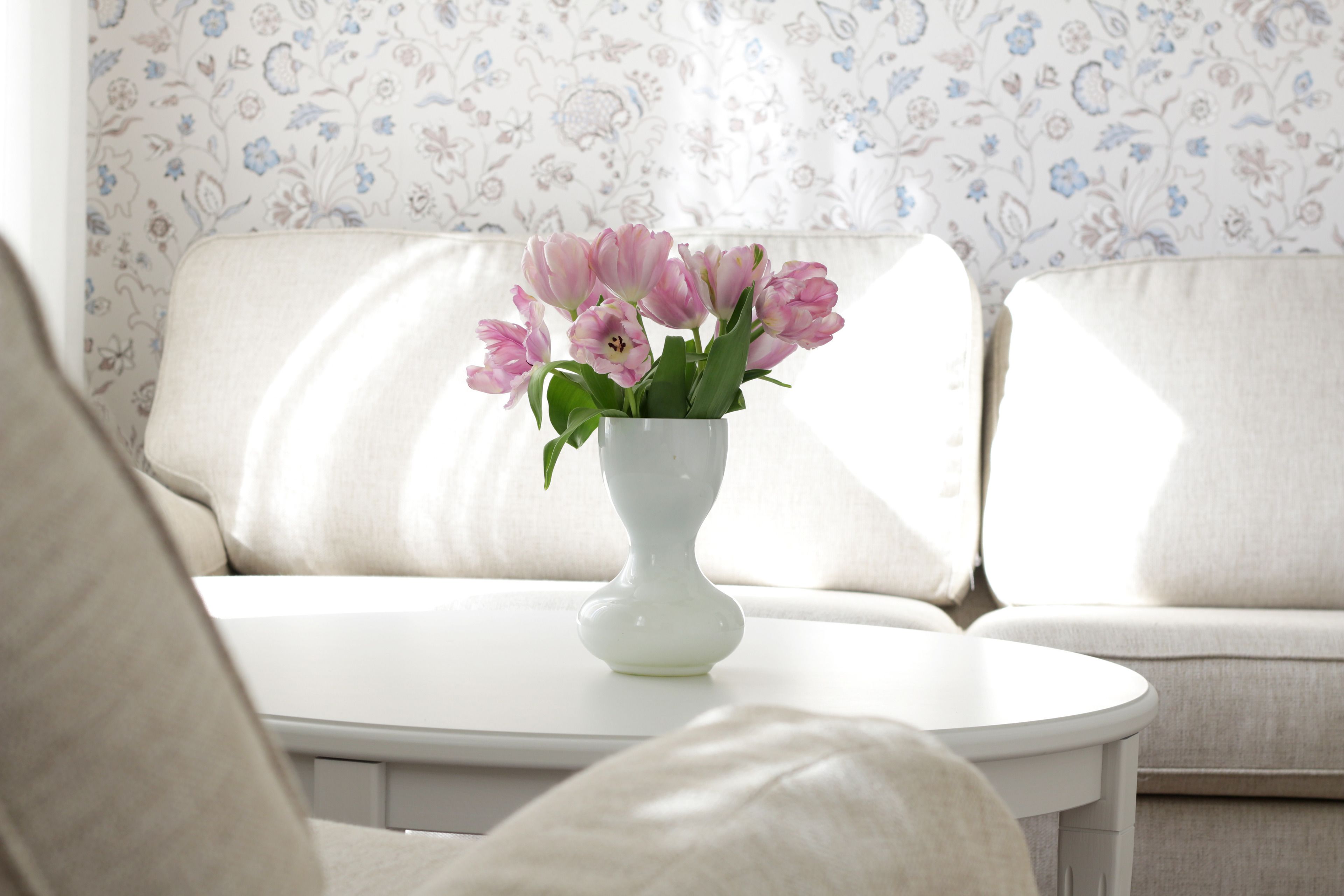 A white vase with pink flowers on a white table in a room with a white sofa and floral wallpaper.