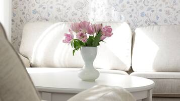 A white vase with pink flowers on a white table in a room with a white sofa and floral wallpaper.