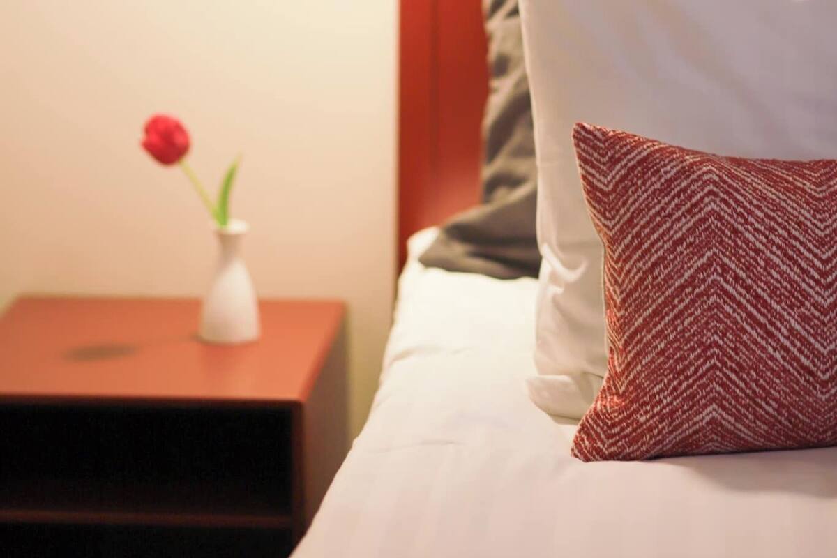 Ein ordentlich bezogenes Bett mit einem rot-weißen Kopfkissen, eine weiße Vase mit einer roten Blume und eine moderne Pendelleuchte.