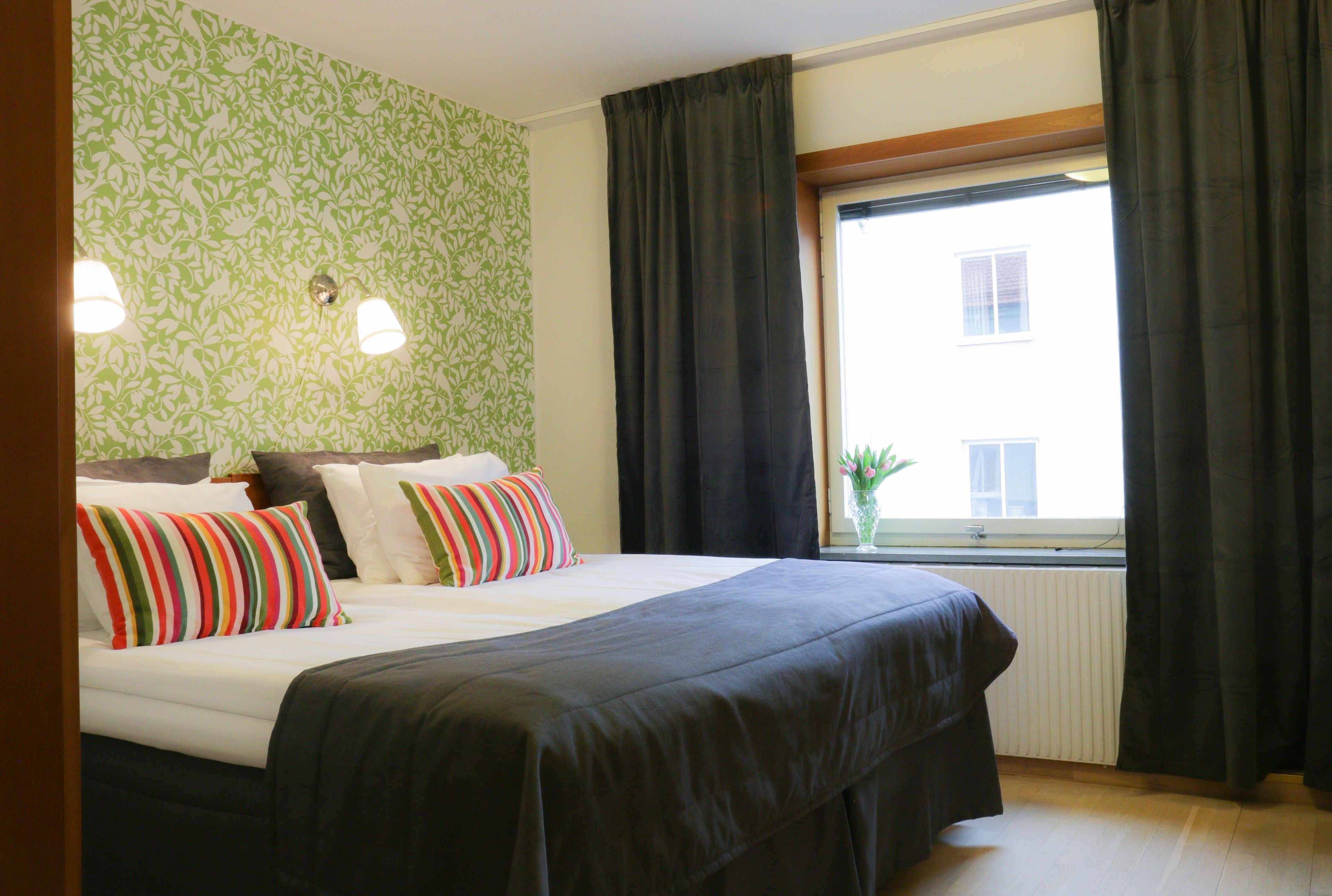 A hotel room with a large bed, two bedside lamps, a window with curtains, and a radiator.