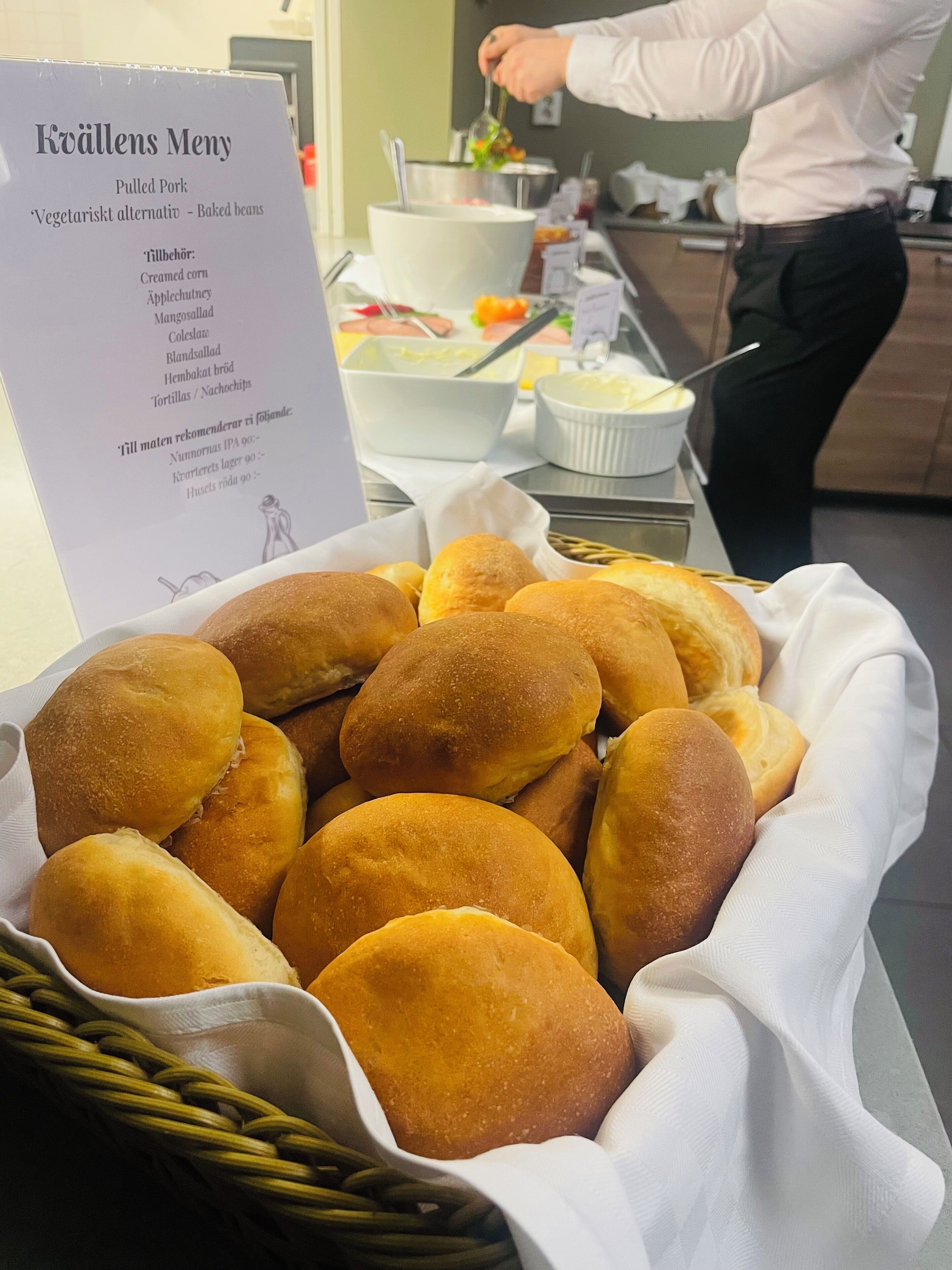 A buffet table with a sign listing pulled pork and vegetarian alternatives, and a basket of golden-brown rolls.