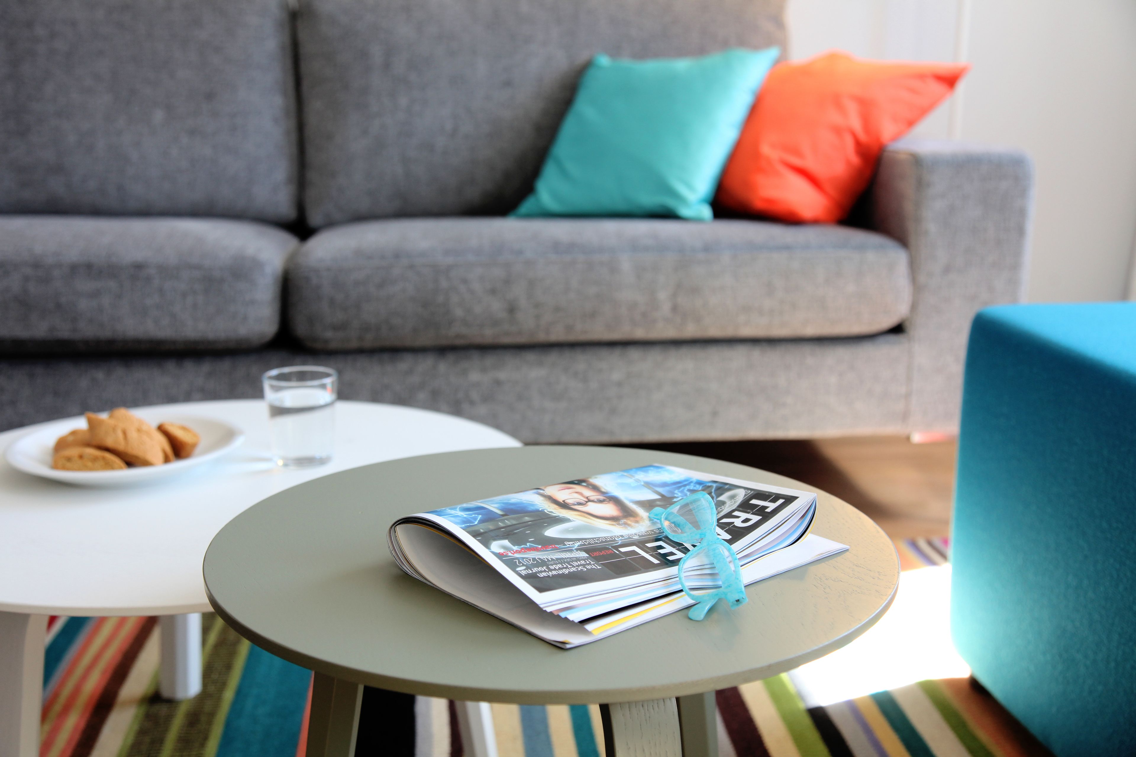 A living room with a grey sofa, a small round table with a magazine and a glass of water, and a plate of cookies.