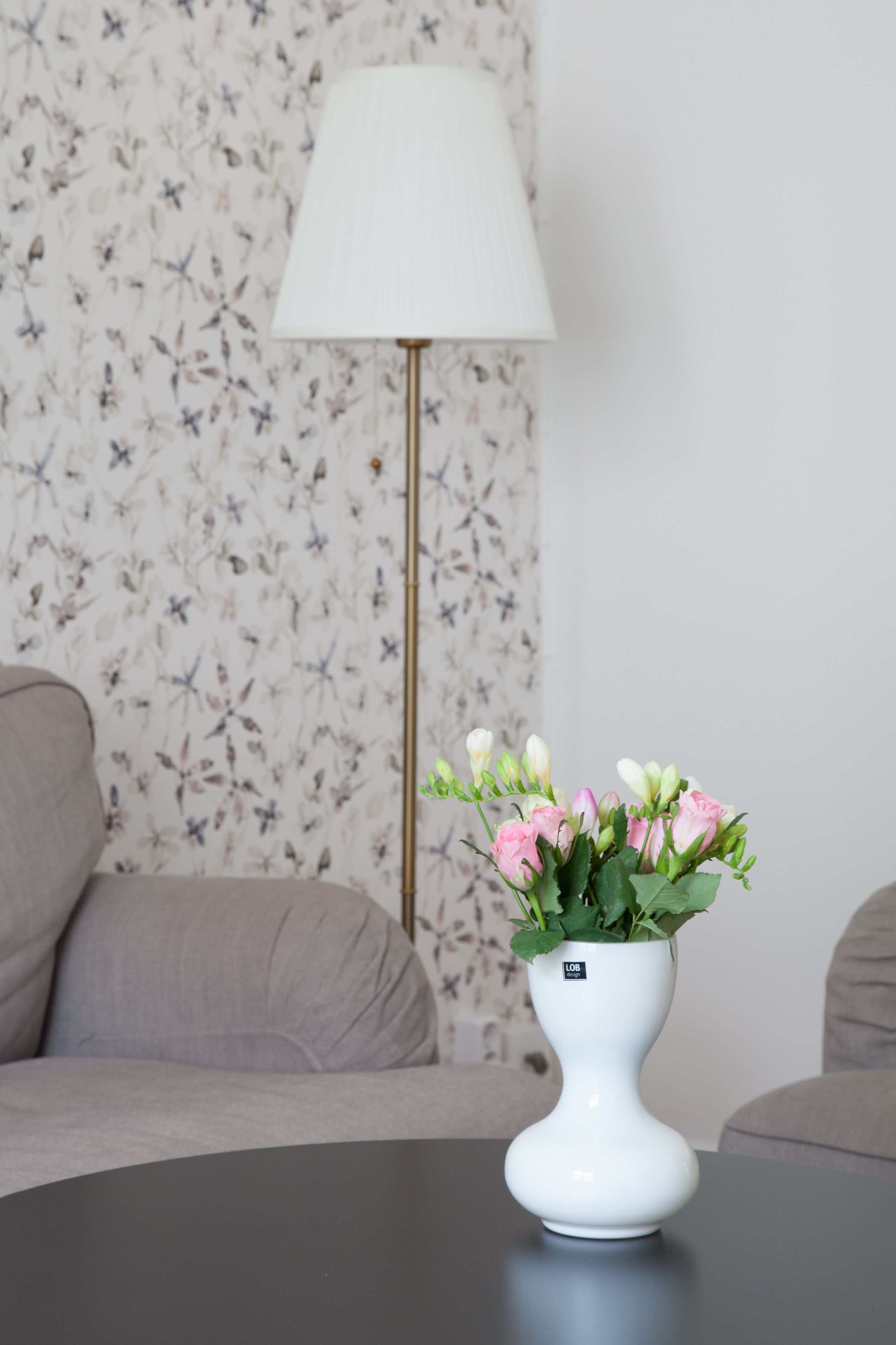 A white vase with pink flowers on a table, a lamp, and a floral wallpaper in the background.