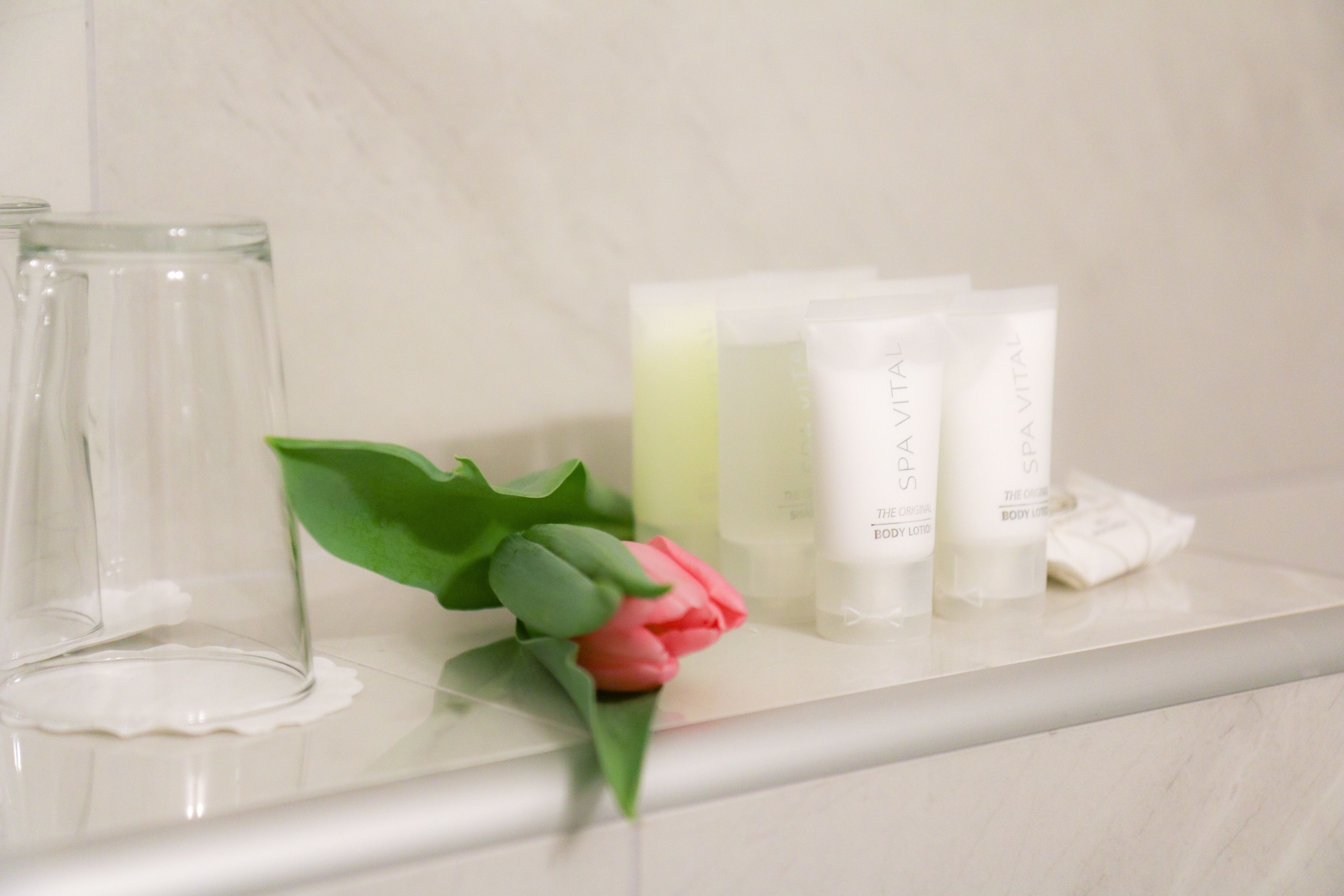 A bathroom shelf with a glass vase, a pink rose, and three bottles of body lotion.