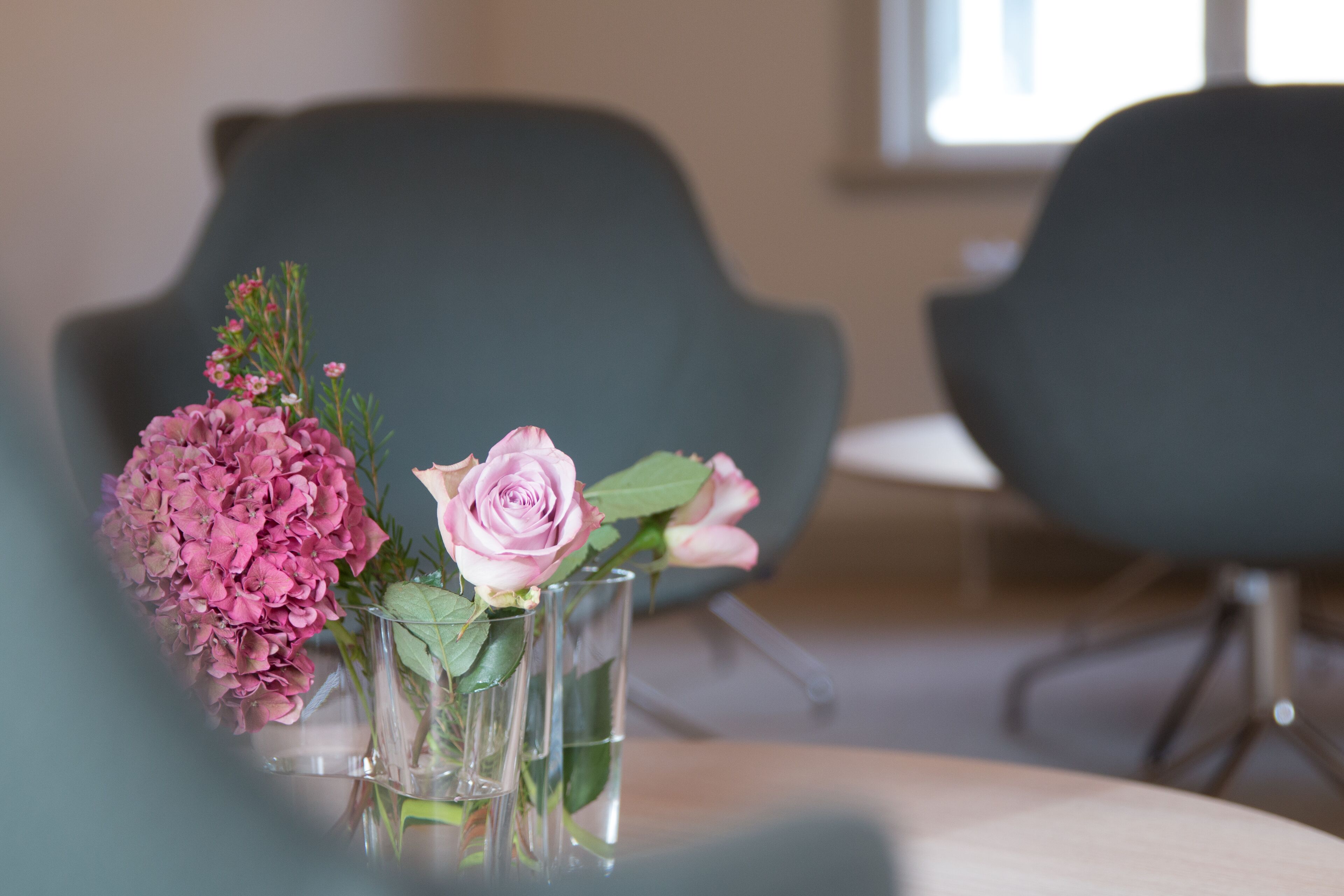 A glass vase with pink flowers on a table in a room with dark chairs.
