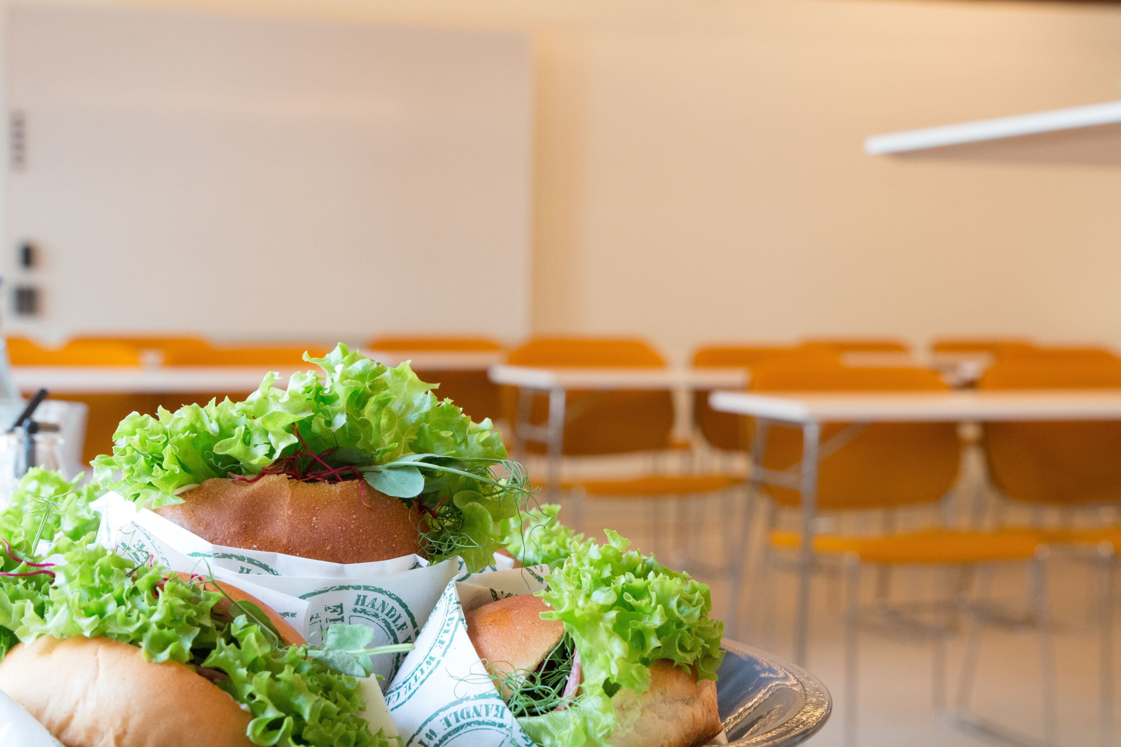 A tray of sandwiches with lettuce and paper on a table in a room with chairs and tables.
