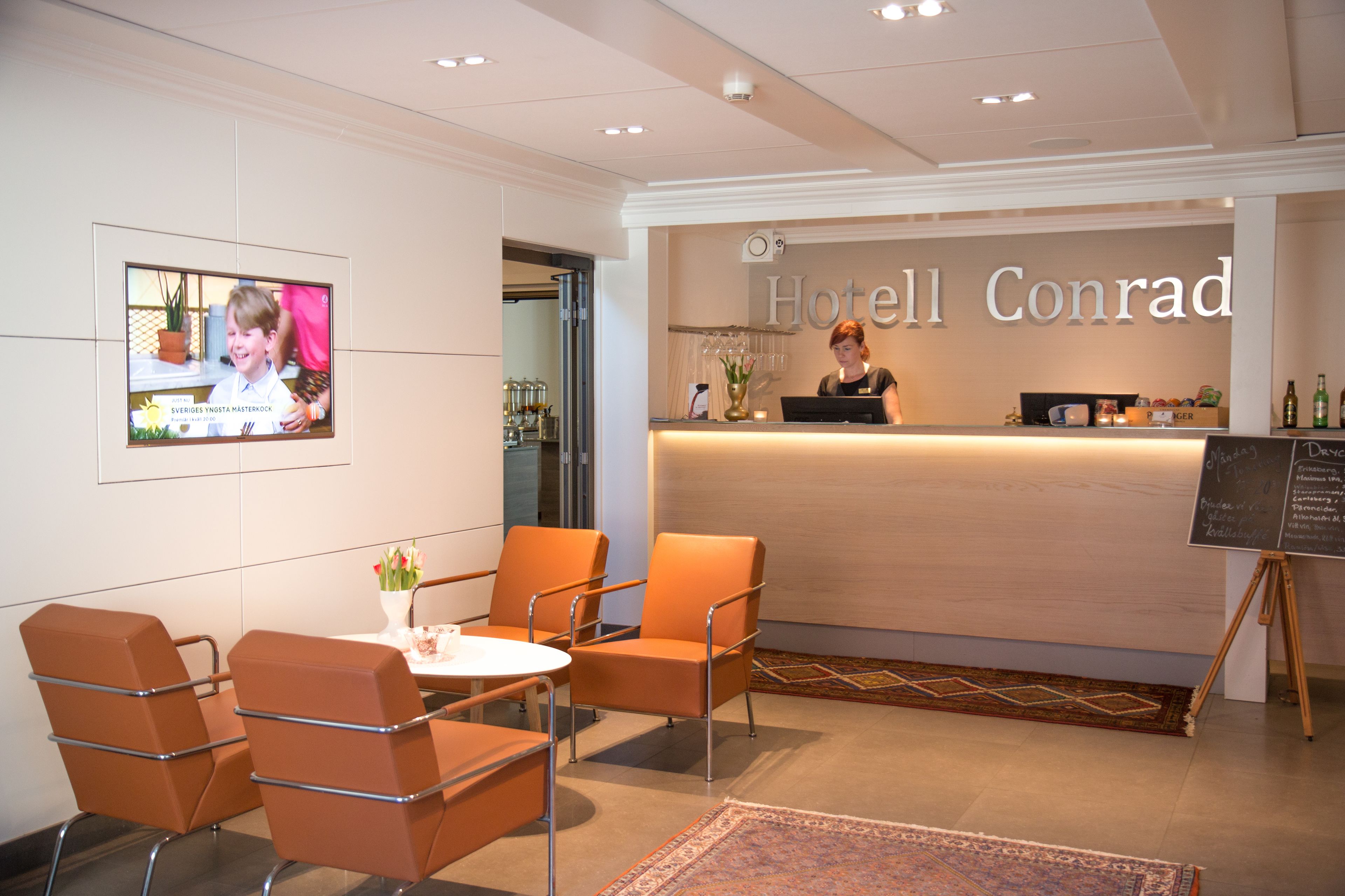 En hotellobby med modern design, som inkluderar en receptionsdisk, en tv-skärm och sittplatser.