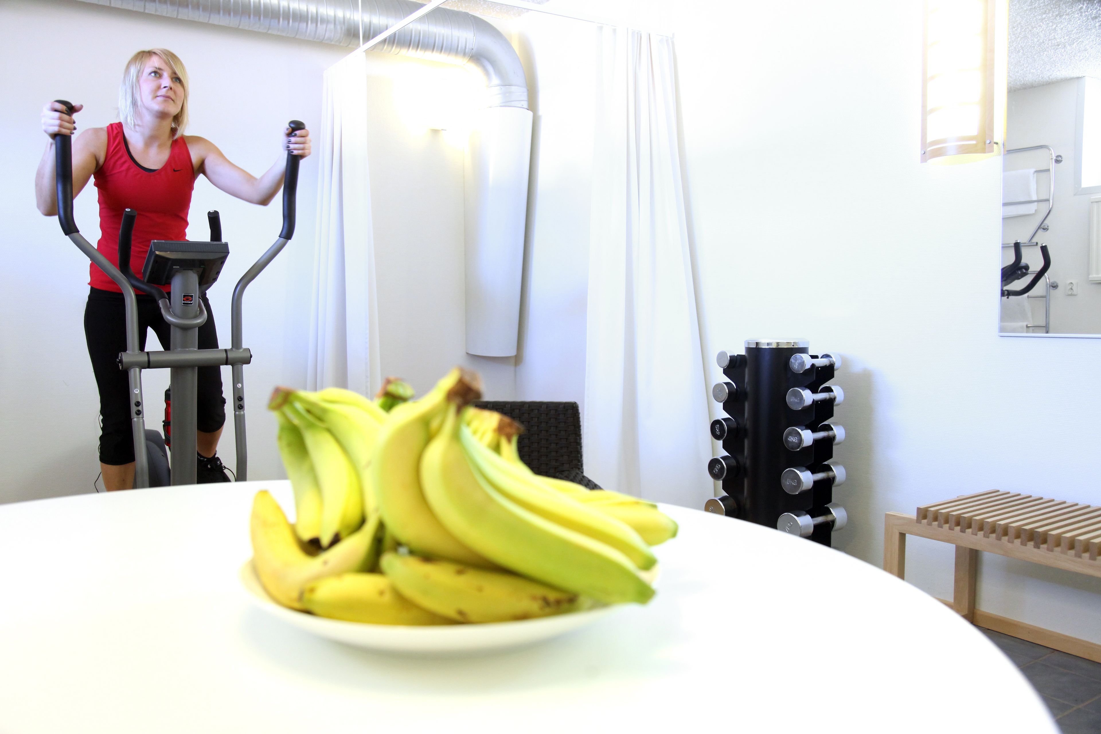 A room with a treadmill, a wall-mounted rack of dumbbells, and a plate of bananas on a table.