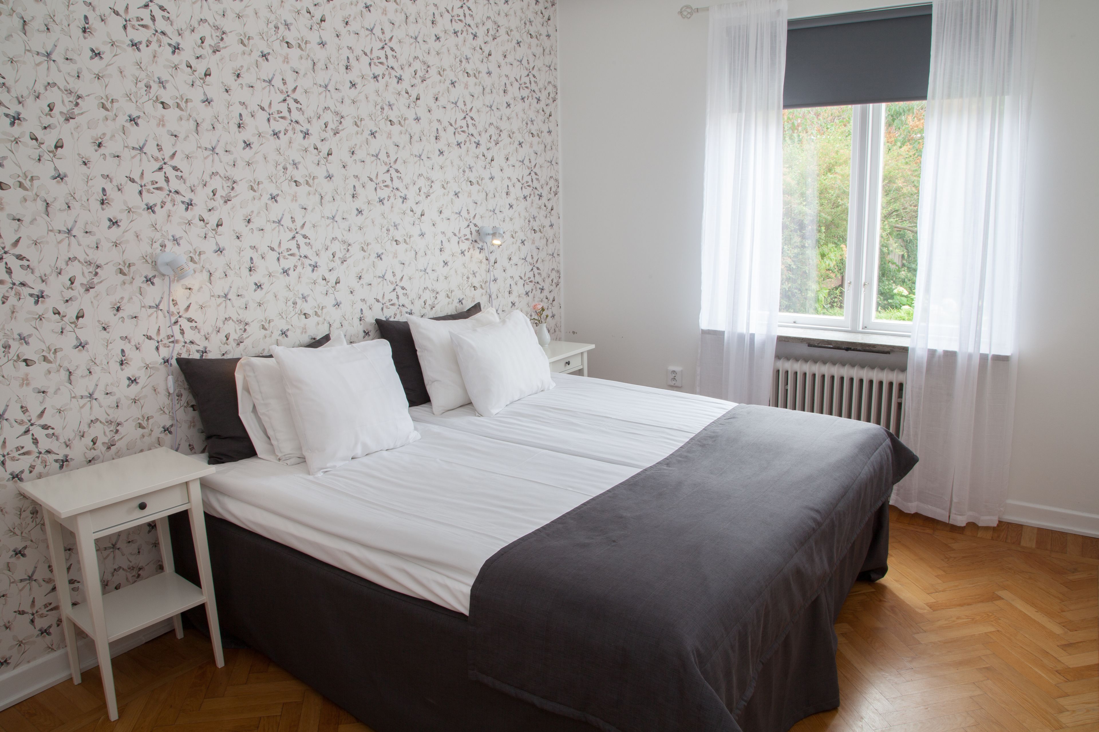 A bedroom with a bed, white nightstands, a small shelf, a window with curtains, and a radiator.