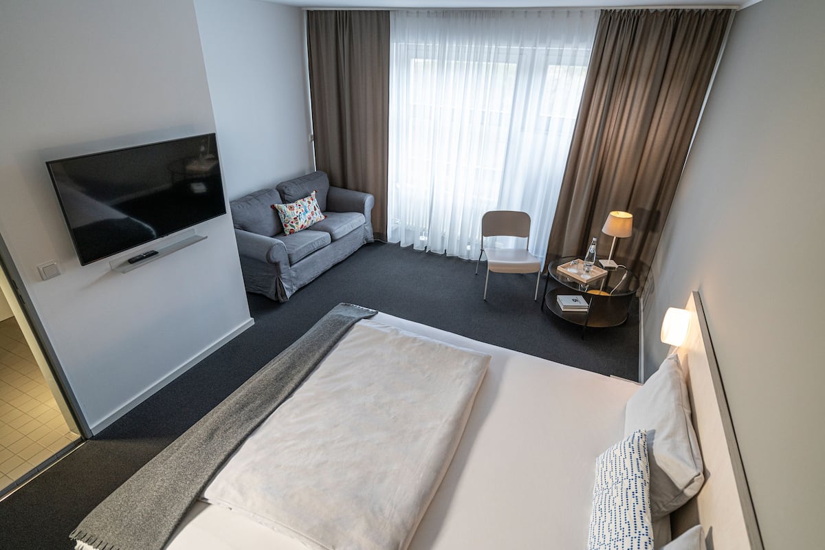 A hotel room with a large bed, a sofa, a TV, a chair, a lamp, and a window with curtains.