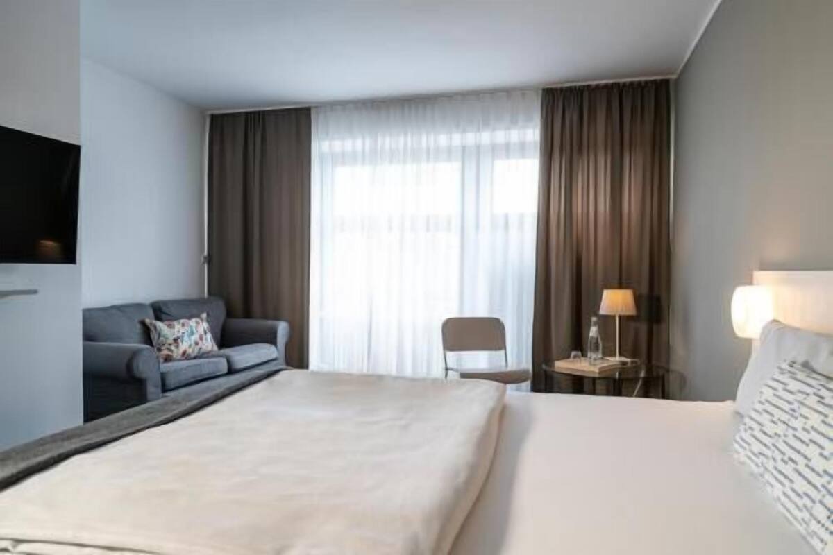 A hotel room with a bed, a sofa, a chair, a TV, and a lamp.