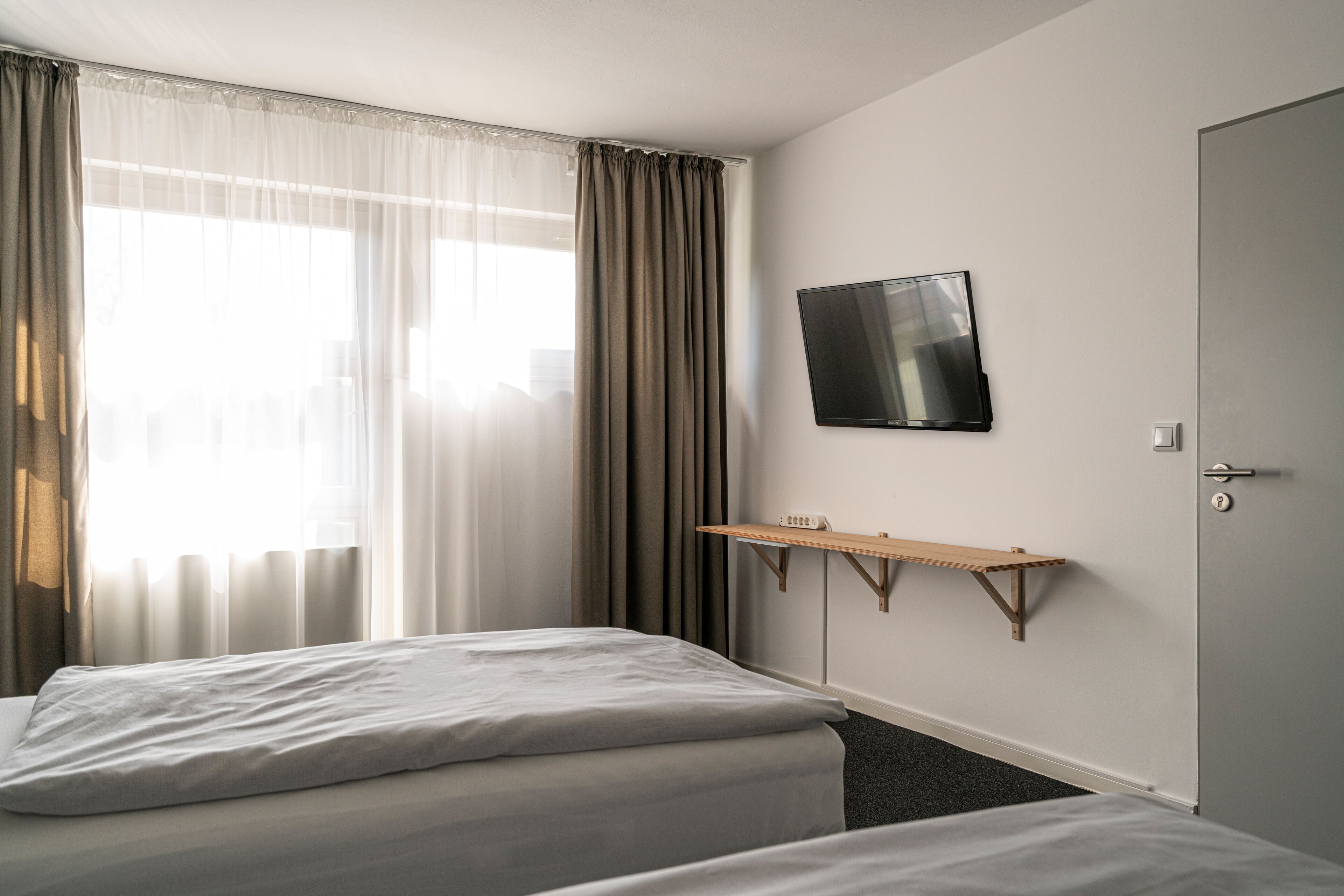 Habitación de hotel con cama, televisor montado en la pared, estante de madera y una puerta.
