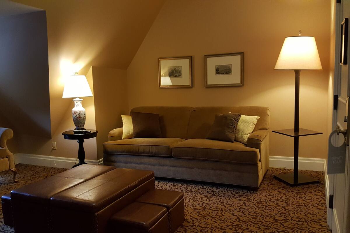 A cozy living room with a brown sofa, two framed pictures on the wall, a side table with a lamp, and a patterned carpet.