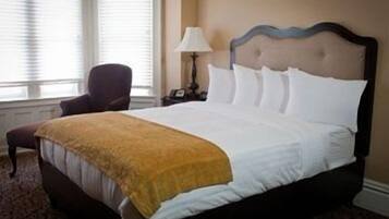 A neatly made bed with white linens and a yellow throw blanket, a dark wooden headboard, and a chair in a room with a window and a nightstand.