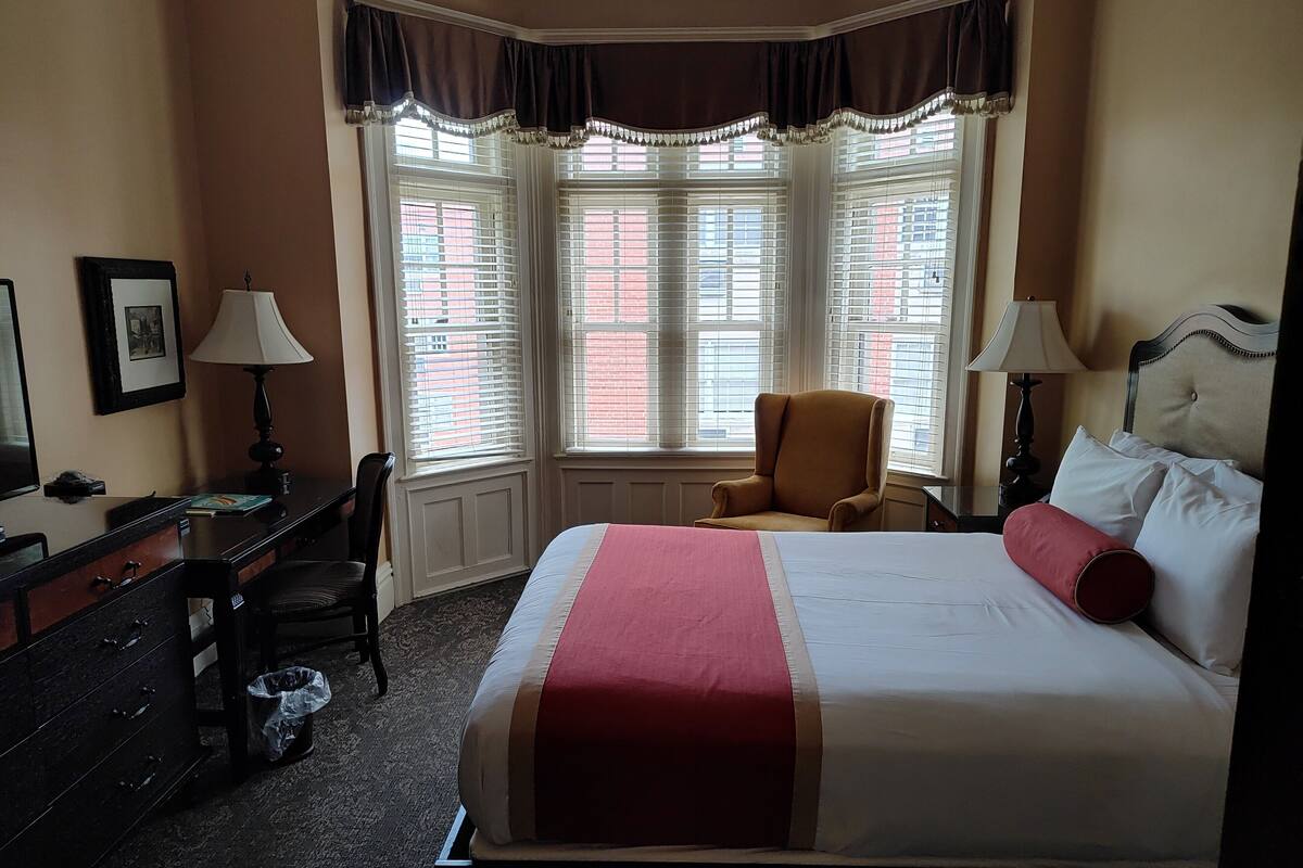 A hotel room with a large bed, a desk, a chair, and a window with blinds.