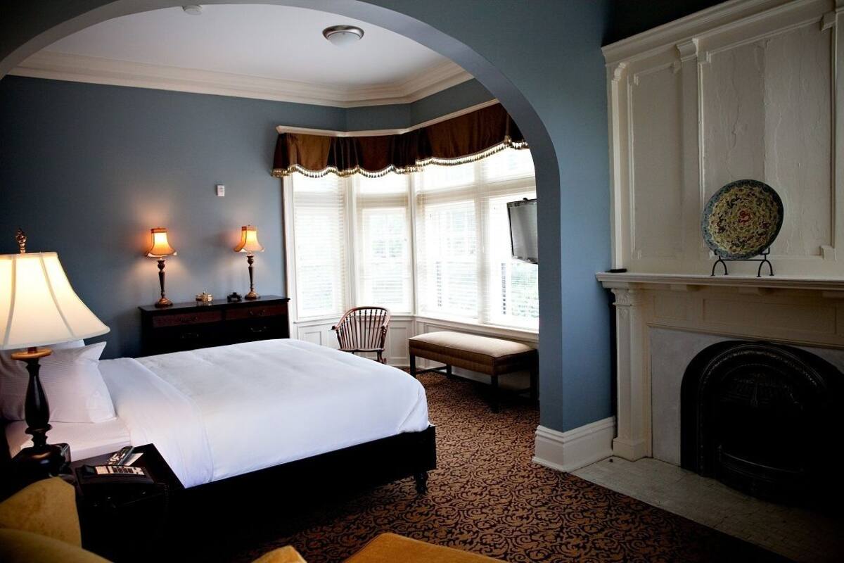 A hotel room with a large bed, a desk with a lamp, a chair, a fireplace, and a window with curtains.