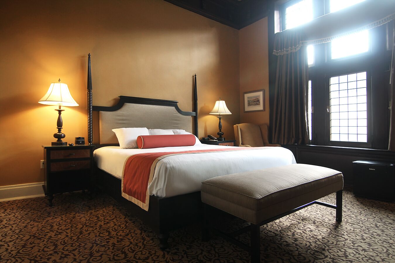 A hotel room with a large bed, two bedside tables with lamps, a chair, a window with curtains, and a patterned carpet.