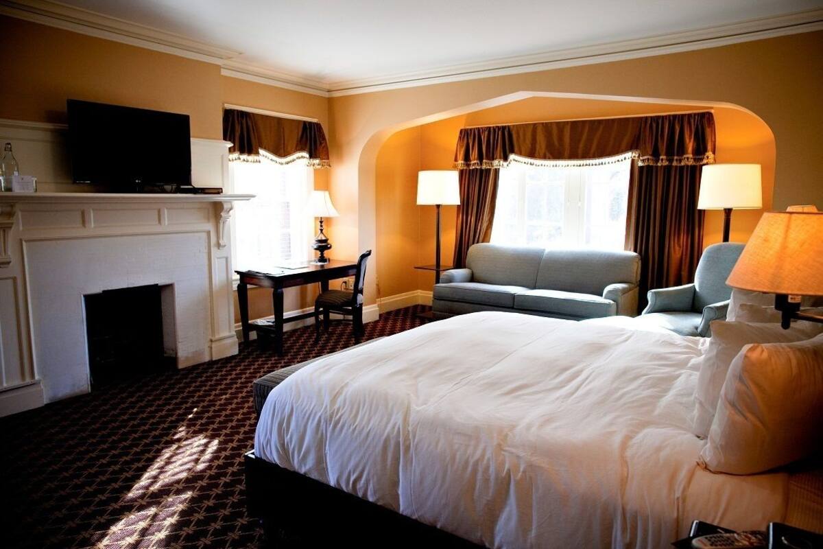 A hotel room with a large bed, a sofa, a desk with a chair, a television, and a fireplace.