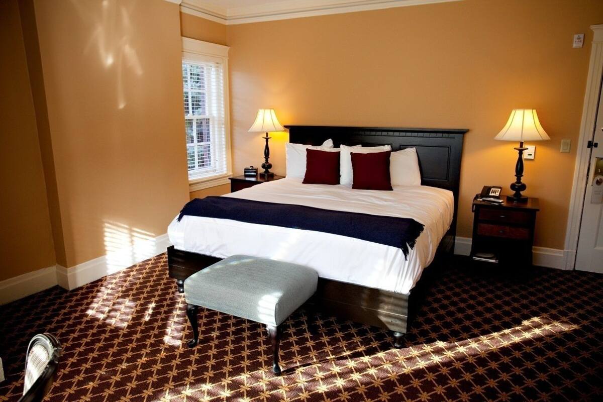 A hotel room with a large bed, two bedside tables with lamps, a window with blinds, and a patterned carpet.