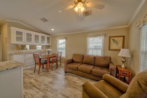 A living room with a brown sofa, a dining table with chairs, and a kitchen area with white cabinets and a ceiling fan. - Arbor Terrace RV Resort (Bradenton)