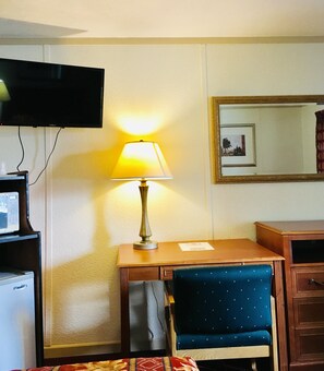 Quarto de hotel com escrivaninha, cadeira, luminária, televisão e espelho.