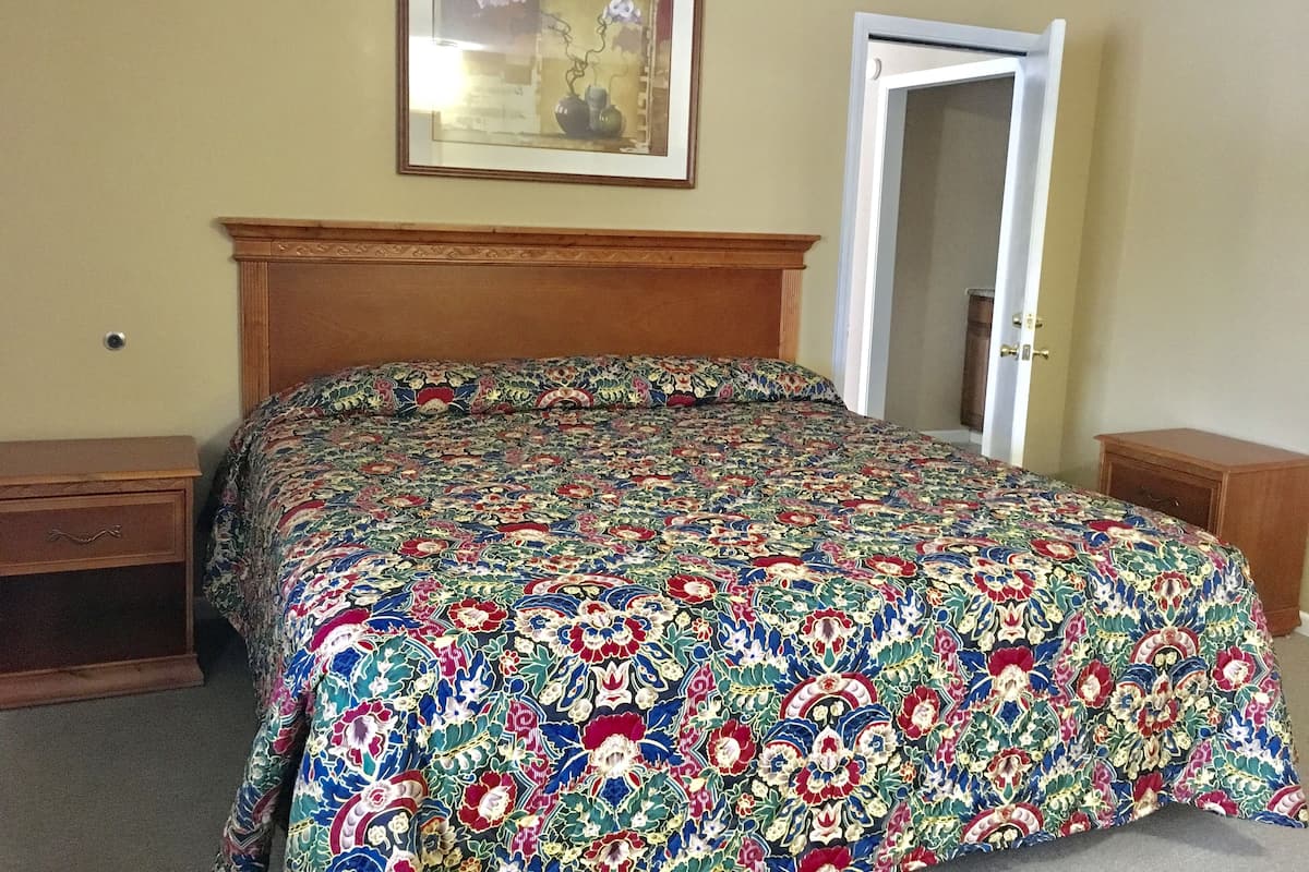 Quarto com cabeceira de madeira, colcha estampada com flores, mesa de cabeceira, gaveta e quadro pendurado na parede.