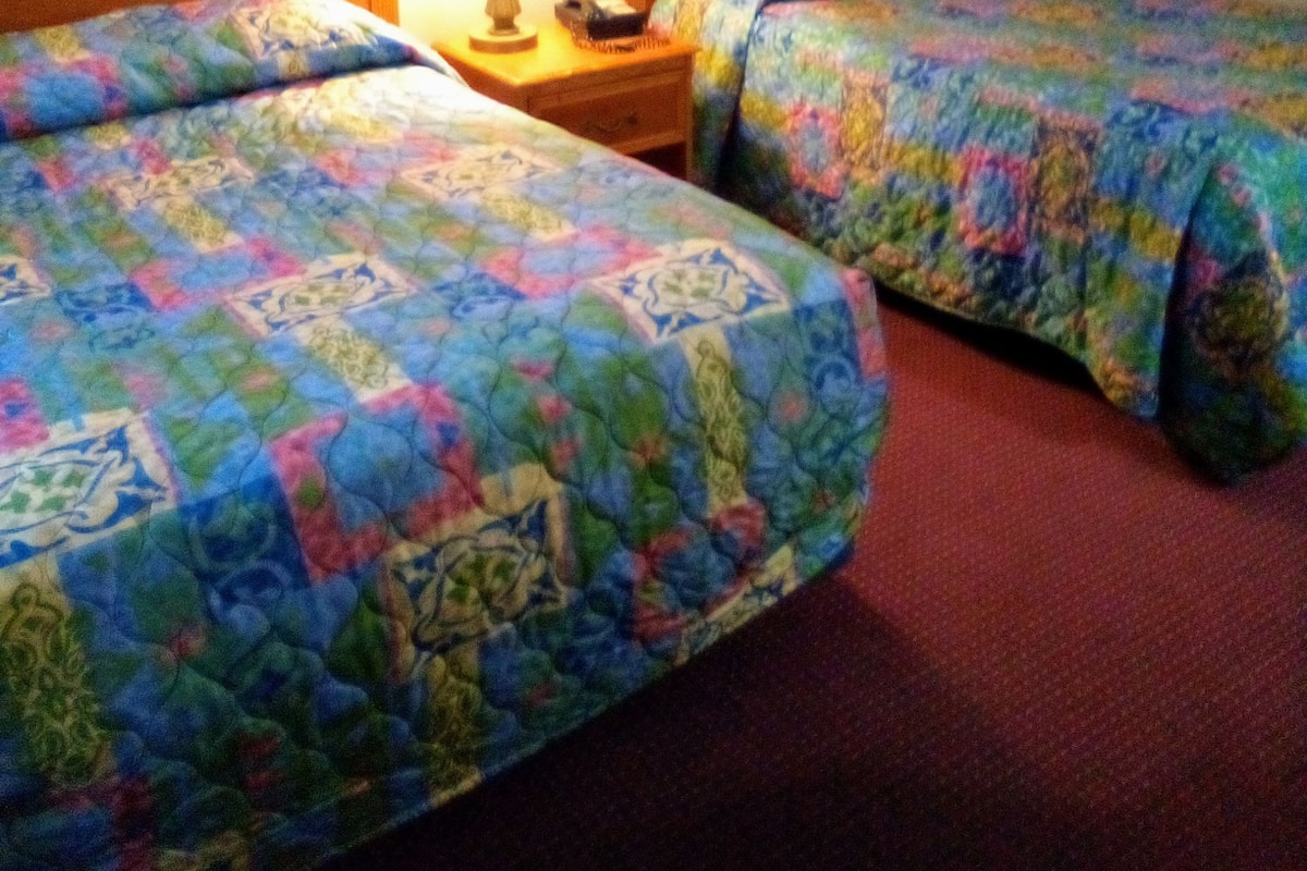 Quarto de hotel com duas camas, cada uma com colcha colorida, luminária de cabeceira e quadros nas paredes.