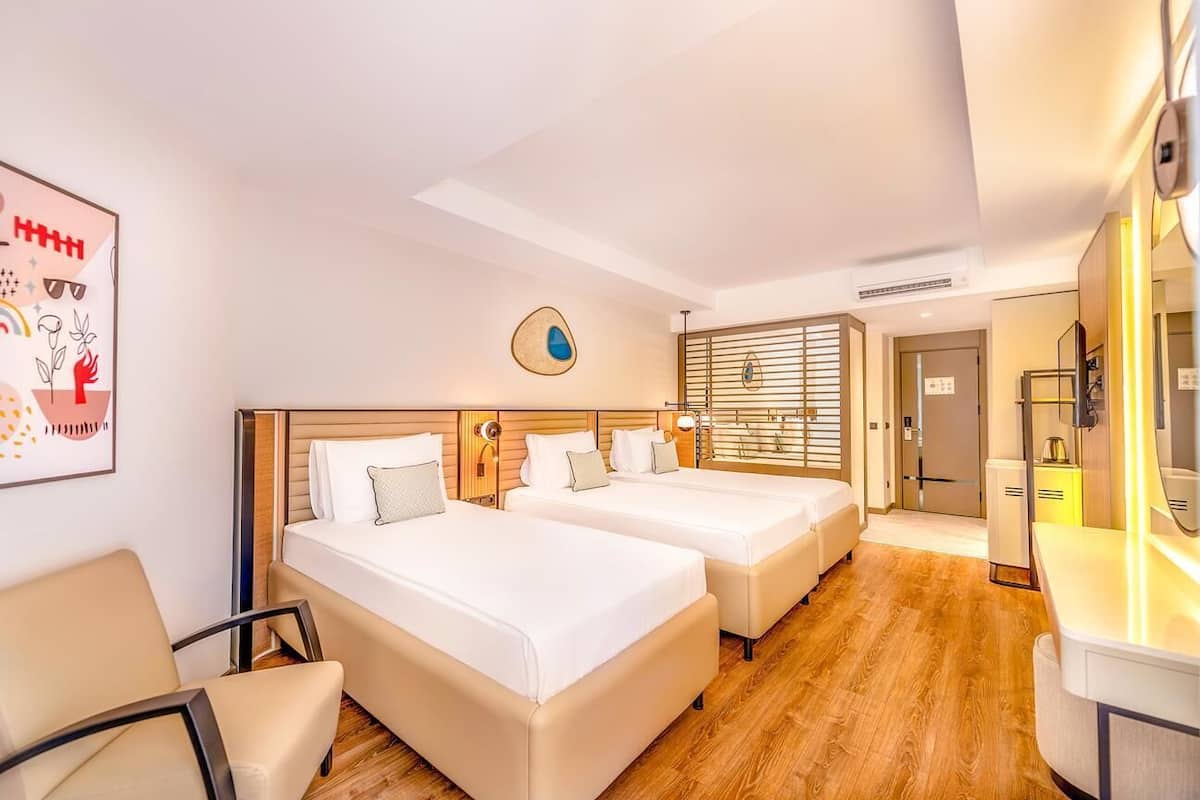 A hotel room with two beds, a desk, a chair, a wall clock, and a window with blinds.