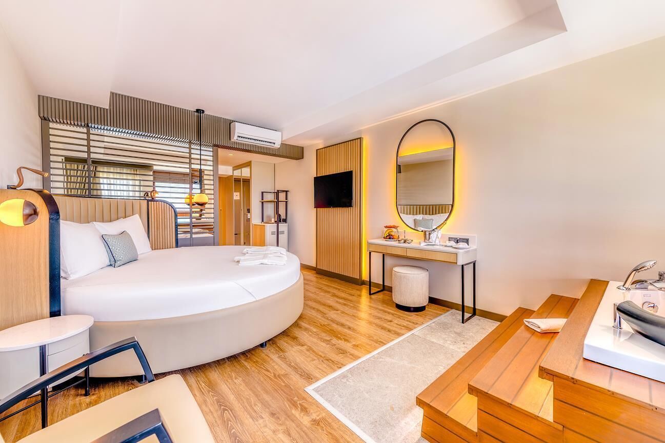 A modern hotel room with a curved bed, a desk, a vanity with a mirror, and a television.