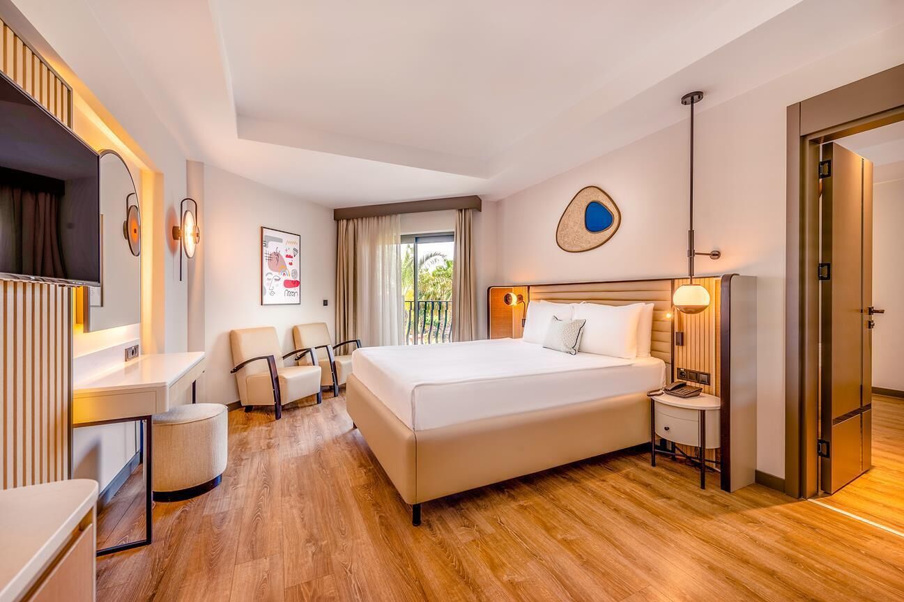 A modern hotel room with a large bed, a desk, a chair, a TV, and a window with curtains.