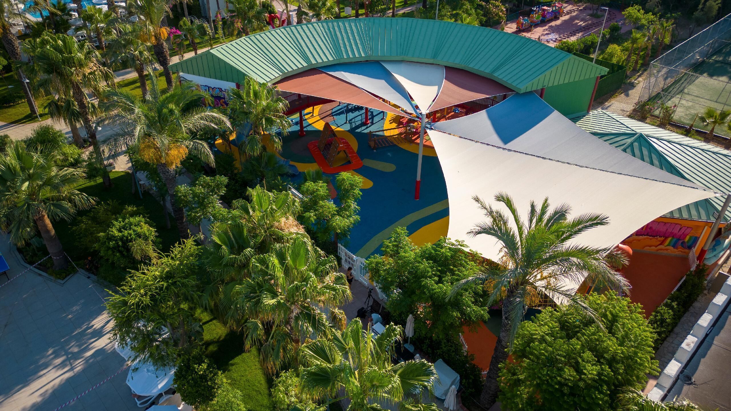 Vista aérea de un resort con un área de juegos colorida, palmeras y un edificio grande con techo curvo.