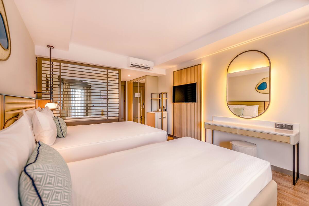 A hotel room with two beds, a desk with a flat-screen TV, a mirror, and a window with blinds.